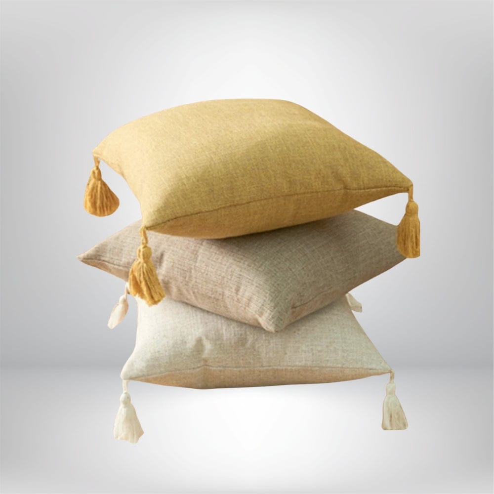 Trois coussins décoratifs aux teintes jaune, beige et crème, avec pompons, posés sur un fond neutre