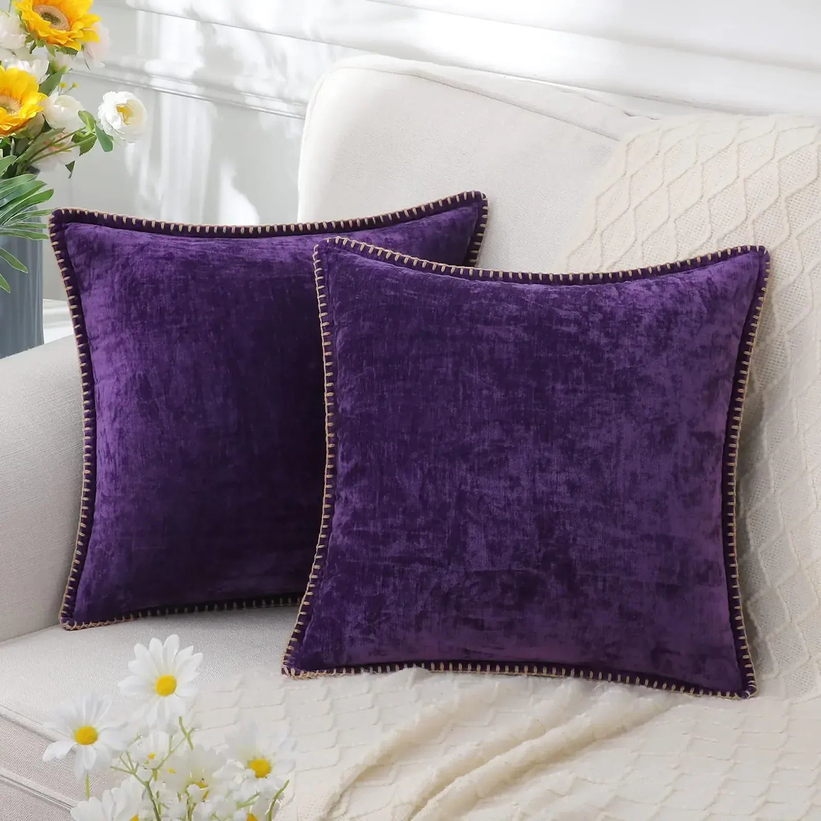 Deux coussins décoratifs en velours violet sur un canapé beige, avec un liseré doré et un plaid en tricot