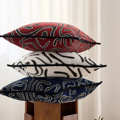 Trois coussins décoratifs aux motifs abstraits rouge, blanc et bleu, posés sur un support en bois
