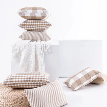 Coussins décoratifs aux motifs à carreaux et rayures, disposés sur un pouf en osier et une couverture blanche