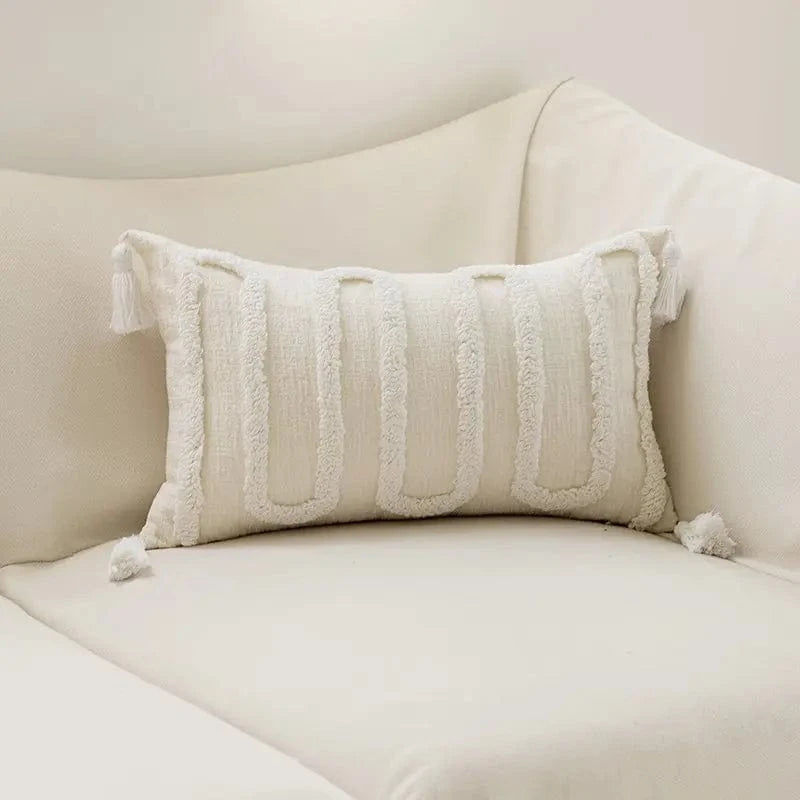 Coussin décoratif blanc à motifs géométriques, posé sur un canapé clair, avec des franges délicates sur les bords