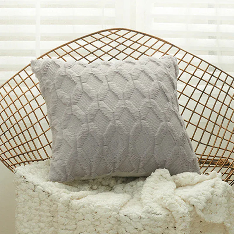 Coussin déco en tissu doux gris avec motif texturé, posé sur un fauteuil en rotin et un plaid blanc