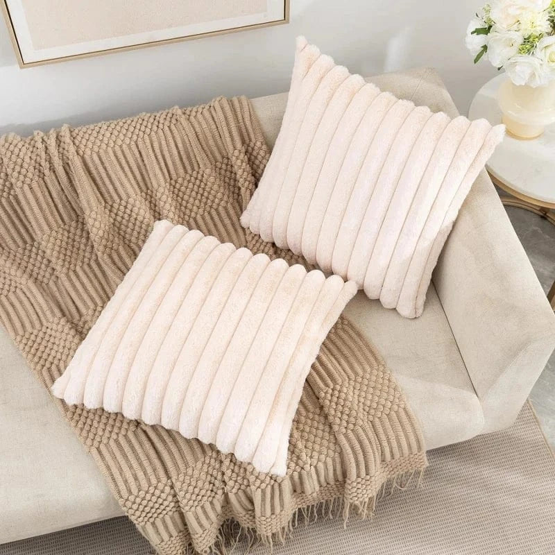 Deux coussins décoratifs en fausse fourrure douce, de couleur rose pâle, posés sur un canapé beige avec un plaid