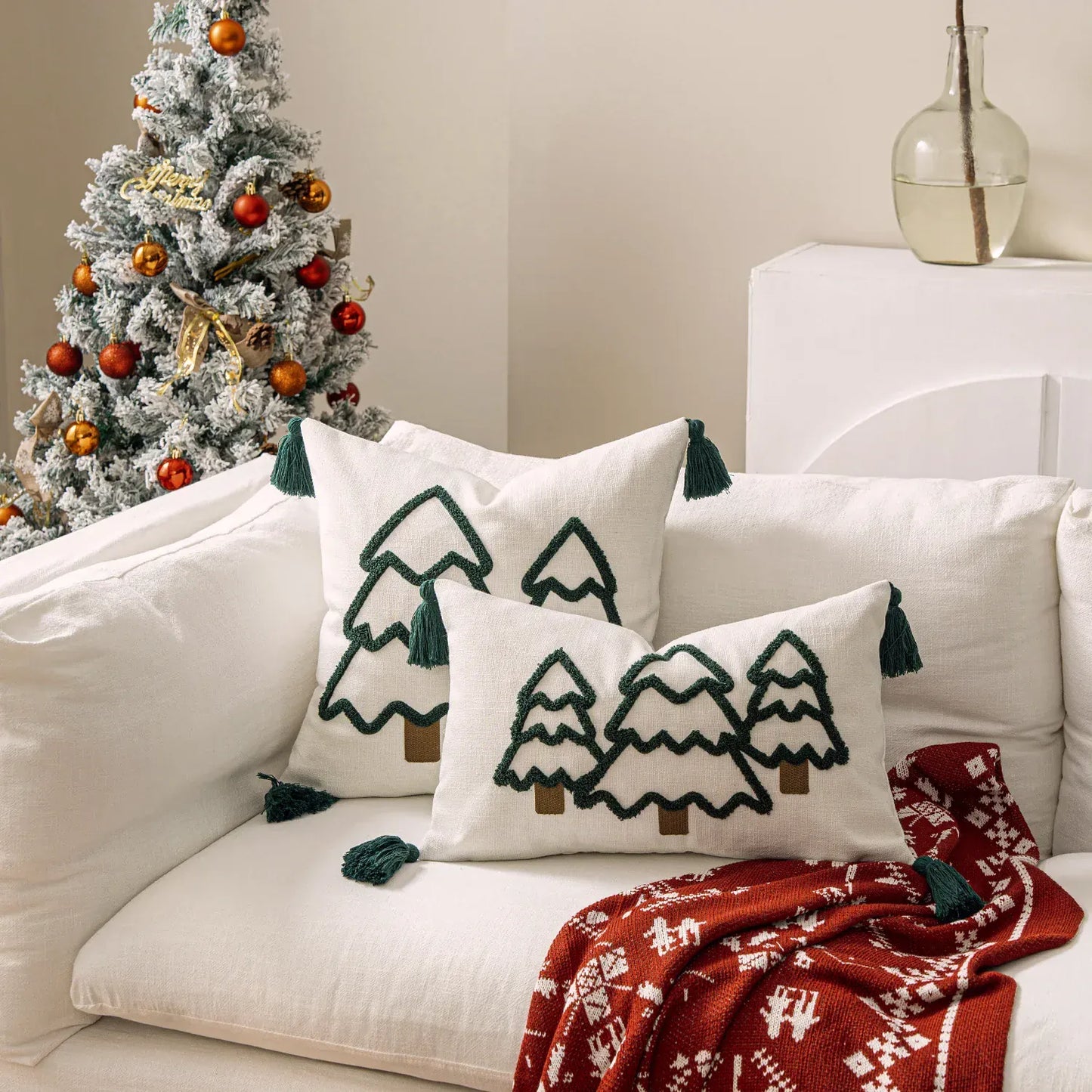 Coussins décoratifs en lin blanc avec motifs de sapins verts et pompons, posés sur un canapé clair