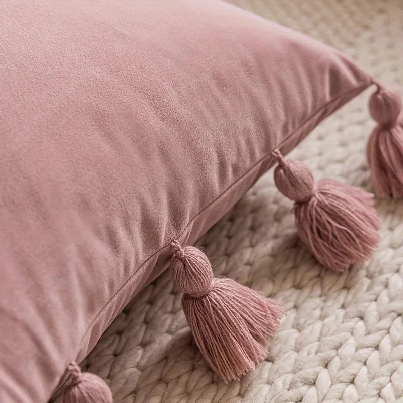 Housse de coussin en velours rose avec des pompons, posée sur un tapis tressé dans un salon chaleureux
