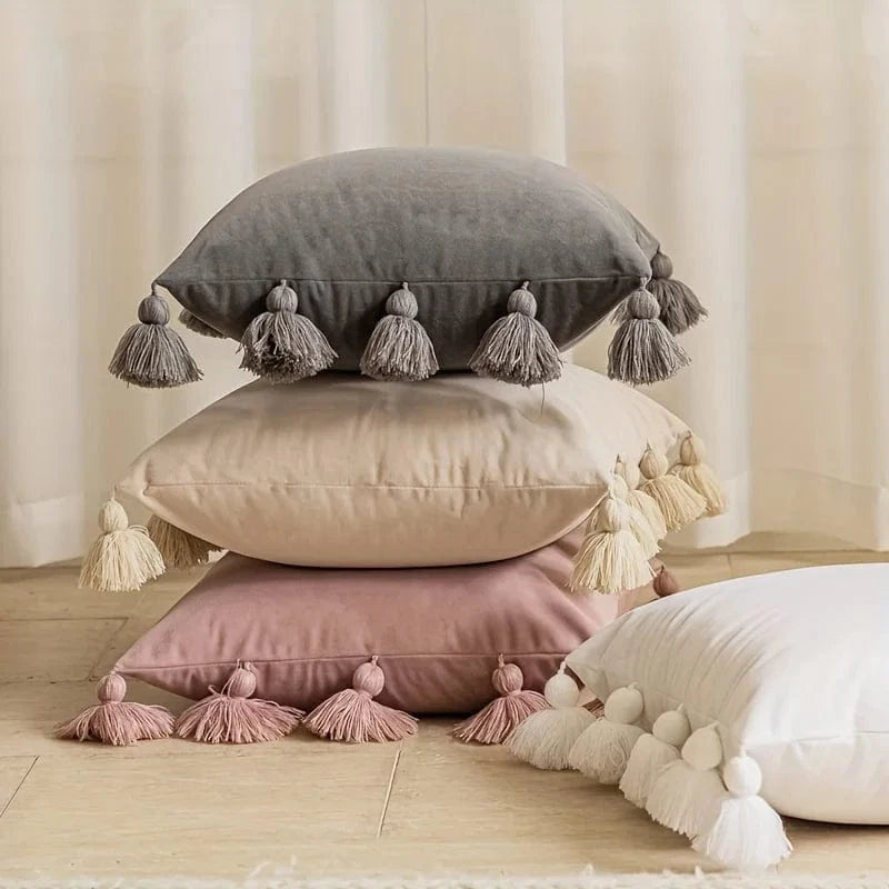 Coussins décoratifs en velours aux teintes douces empilés sur un sol en bois, ornés de pompons colorés