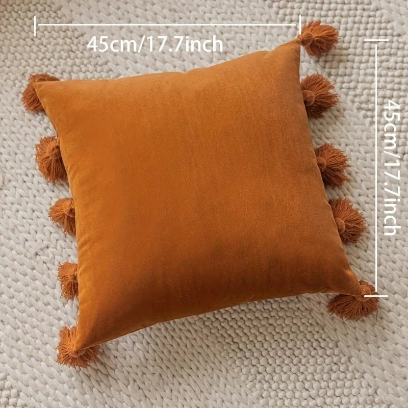 Coussin déco en velours orange avec pompons, posé sur un tapis tissé dans un salon chaleureux