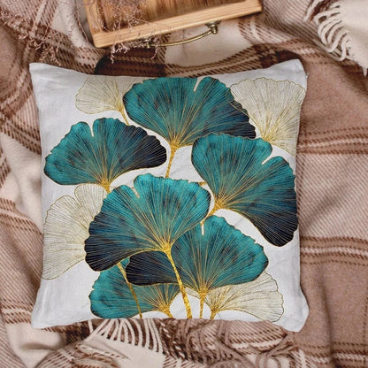 Coussin décoratif avec motifs de feuilles turquoise et dorées, posé sur un plaid à carreaux dans un salon