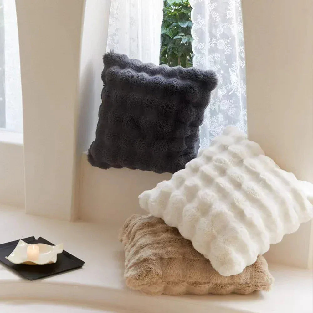 Trois coussins décoratifs en fourrure, de couleurs gris, blanc et beige, posés sur un banc près d'une fenêtre