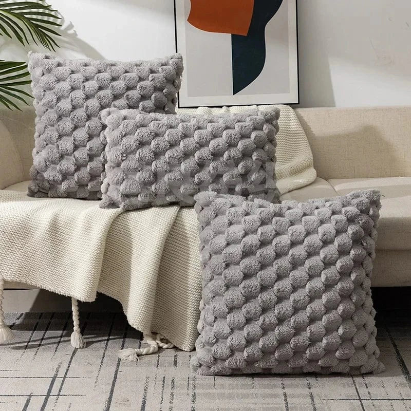 Trois coussins décoratifs gris en matière douce avec motifs en relief, posés sur un canapé avec un plaid