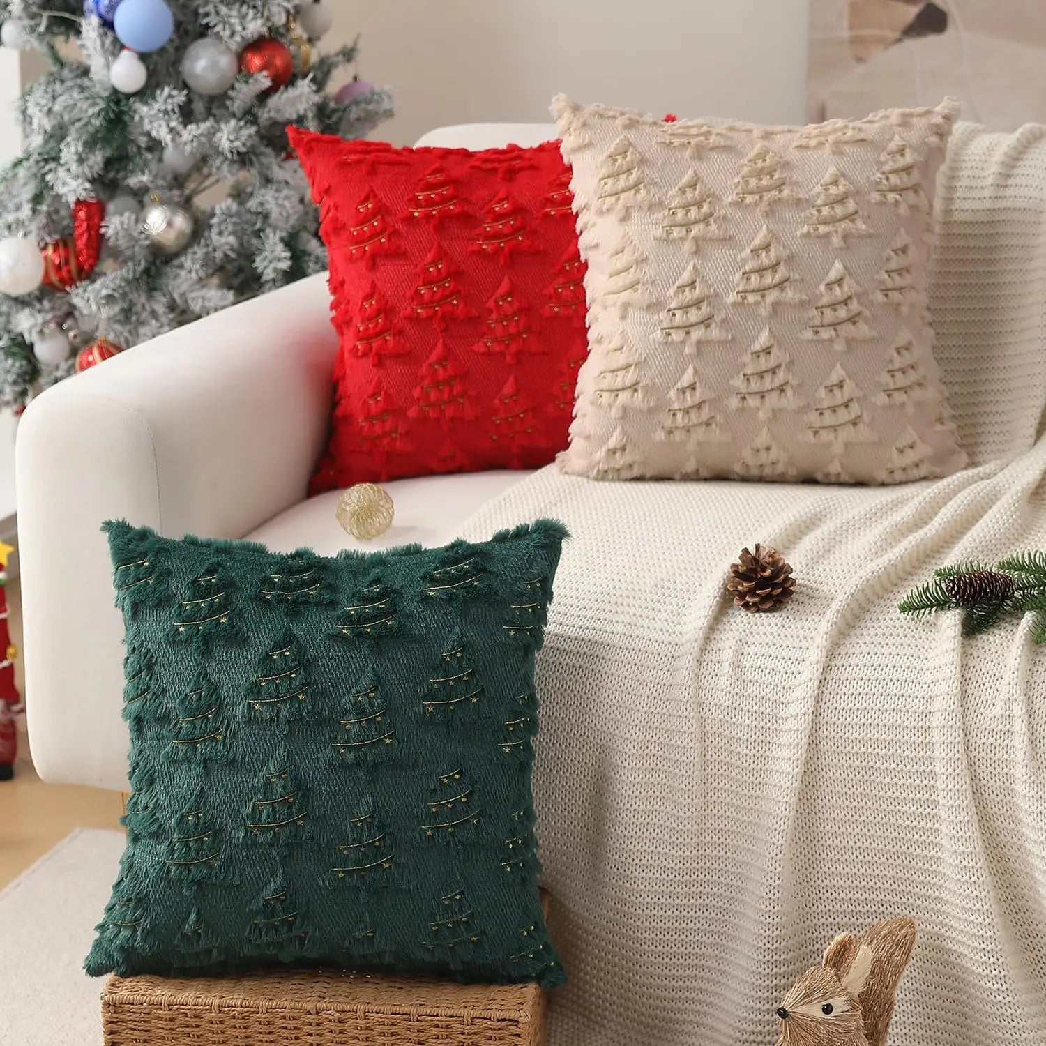 Coussins décoratifs en velours vert, rouge et beige avec motifs de sapins, posés sur un canapé blanc
