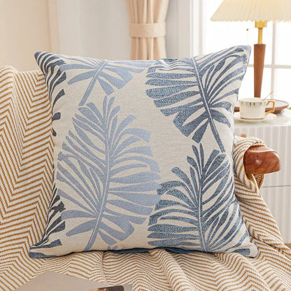 Coussin décoratif aux motifs de feuilles tropicales bleues sur fond beige, posé sur un canapé à rayures