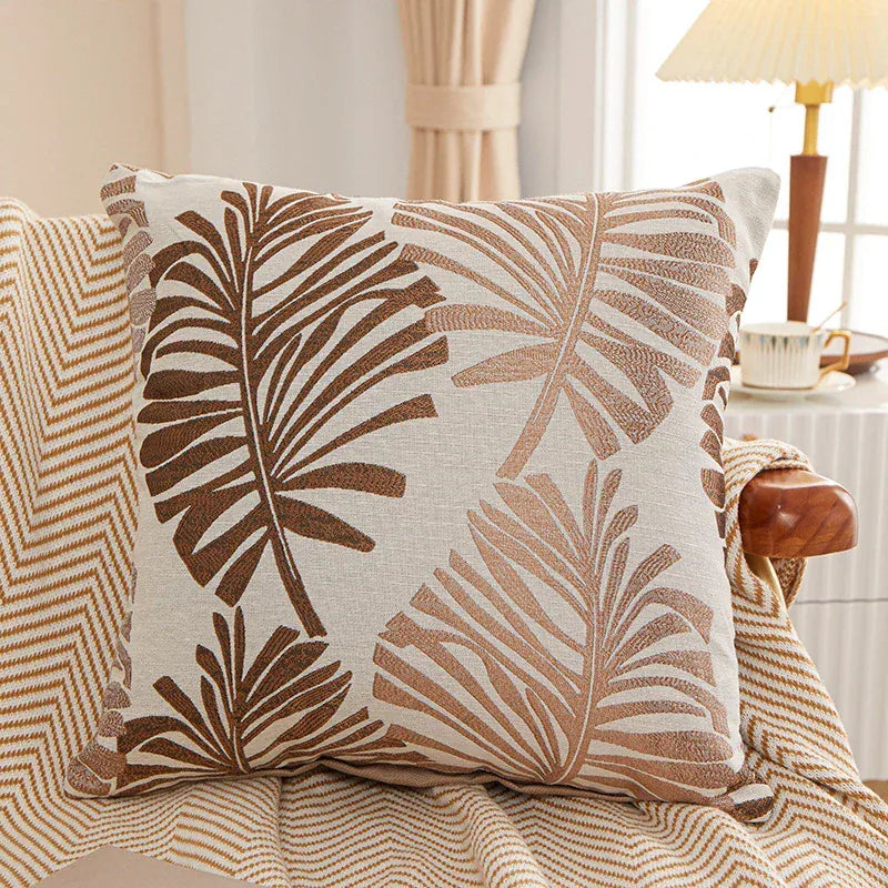 Coussin décoratif aux motifs de feuilles tropicales brunes sur fond beige, posé sur un canapé avec un plaid