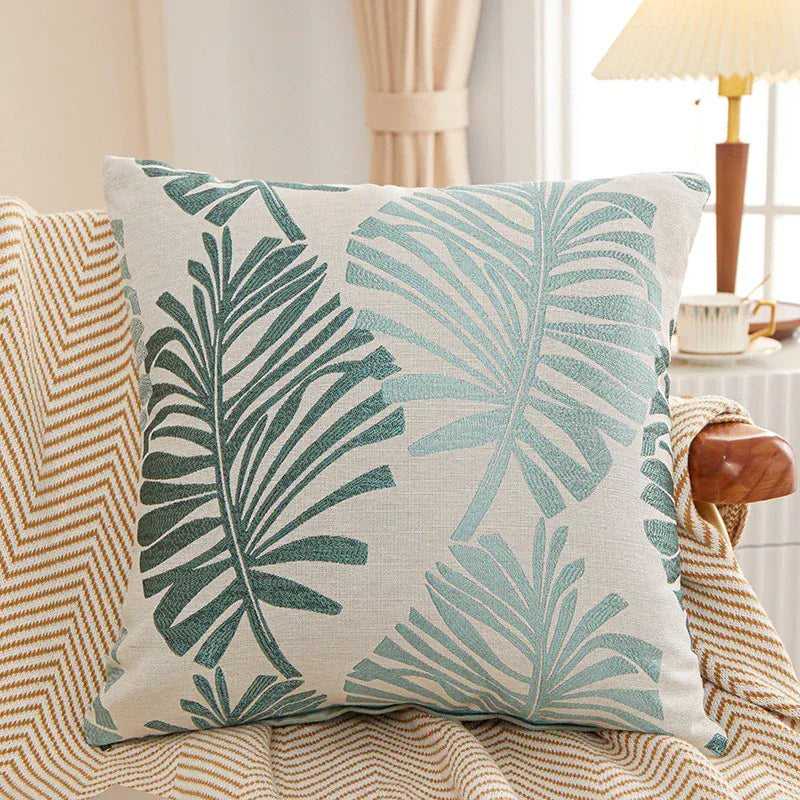 Coussin décoratif à motifs de feuilles tropicales vertes sur fond beige, posé sur un canapé en tissu