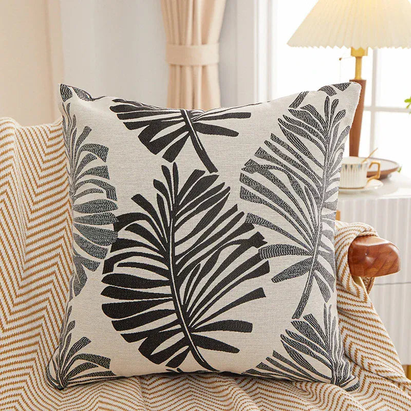 Housse de coussin décoratif avec motifs de feuilles noires sur fond beige, posée sur un canapé