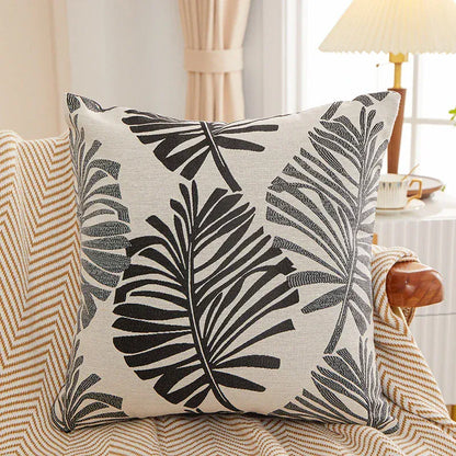 Housse de coussin décoratif avec motifs de feuilles noires sur fond beige, posée sur un canapé