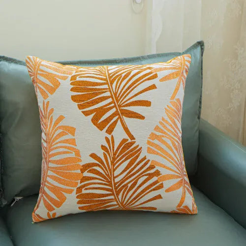 Coussin décoratif aux motifs de feuilles tropicales orange sur fond crème, posé sur un canapé en cuir
