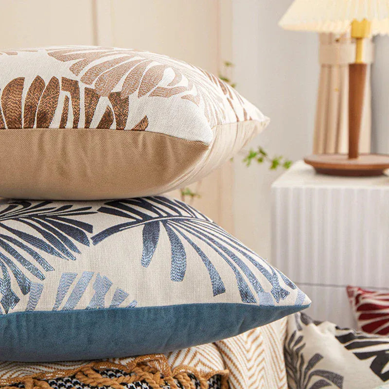 Coussins décoratifs aux motifs tropicaux, avec des couleurs beige, bleu et des feuilles stylisées, sur un canapé