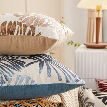 Coussins décoratifs aux motifs tropicaux, avec des couleurs beige, bleu et des feuilles stylisées, sur un canapé